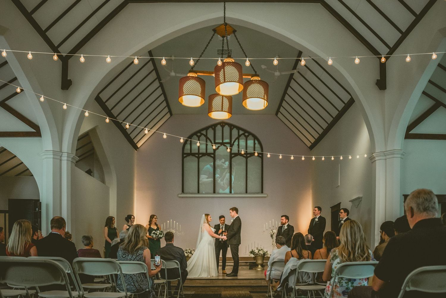 Wedding ceremony taking place in a historic church with vaulted ceilings and string lights creating a romantic atmosphere.