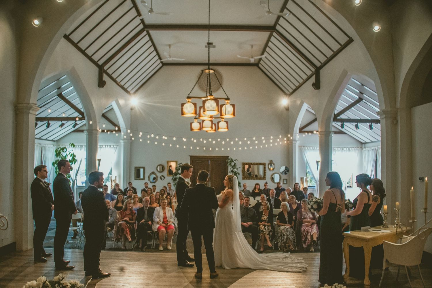 Indoor wedding ceremony in a historic chapel with vaulted ceilings, string lights, and a grand chandelier illuminating the space.