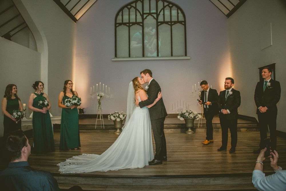 Wedding party stands in a line during a ceremony in a historic chapel with arched windows and elegant lighting.