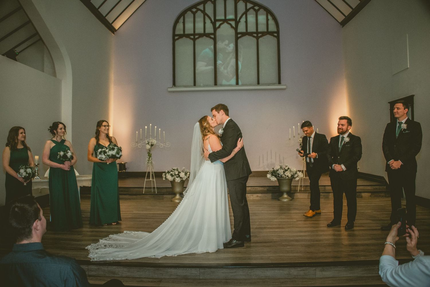 Wedding party stands in a line during a ceremony in a historic chapel with arched windows and elegant lighting.