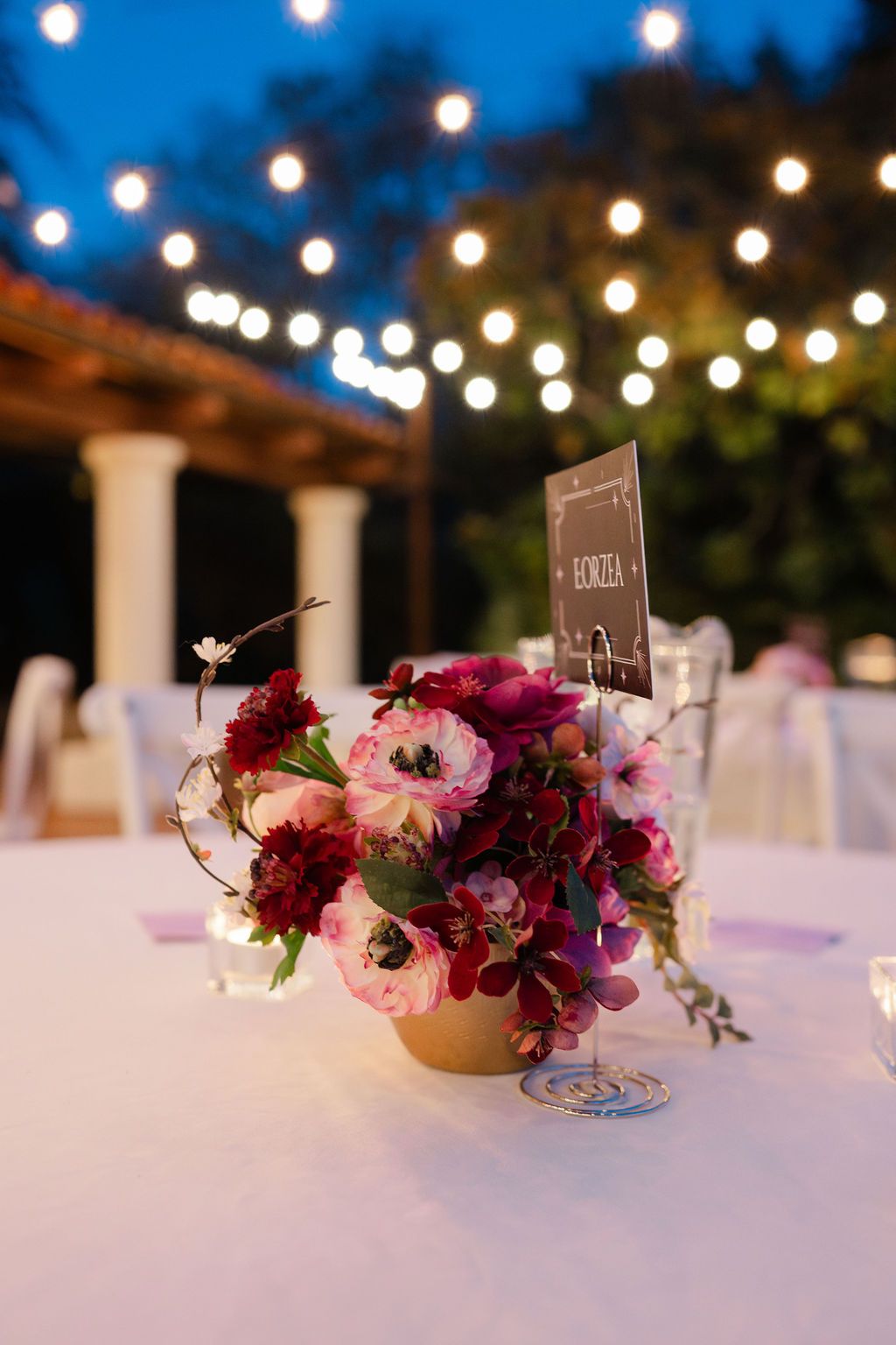 String lights illuminate a wedding reception table with pink and burgundy floral centerpieces on white linens at dusk.