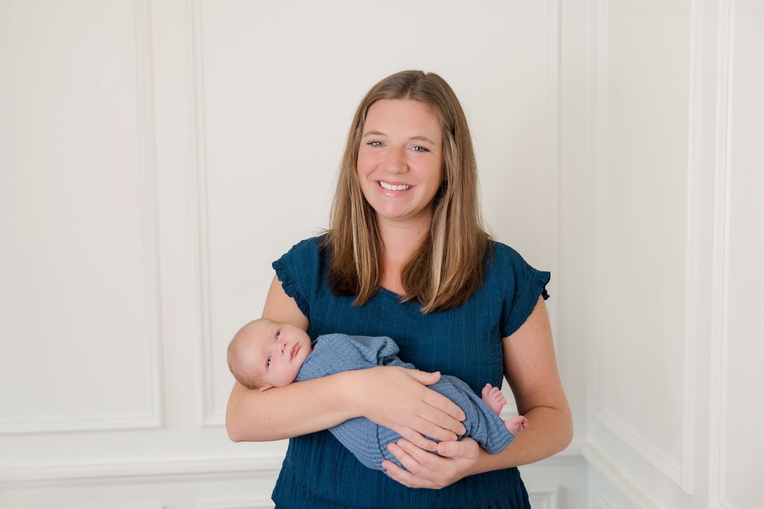 A person wearing a blue shirt holds and plays with a small baby against a white wall background.