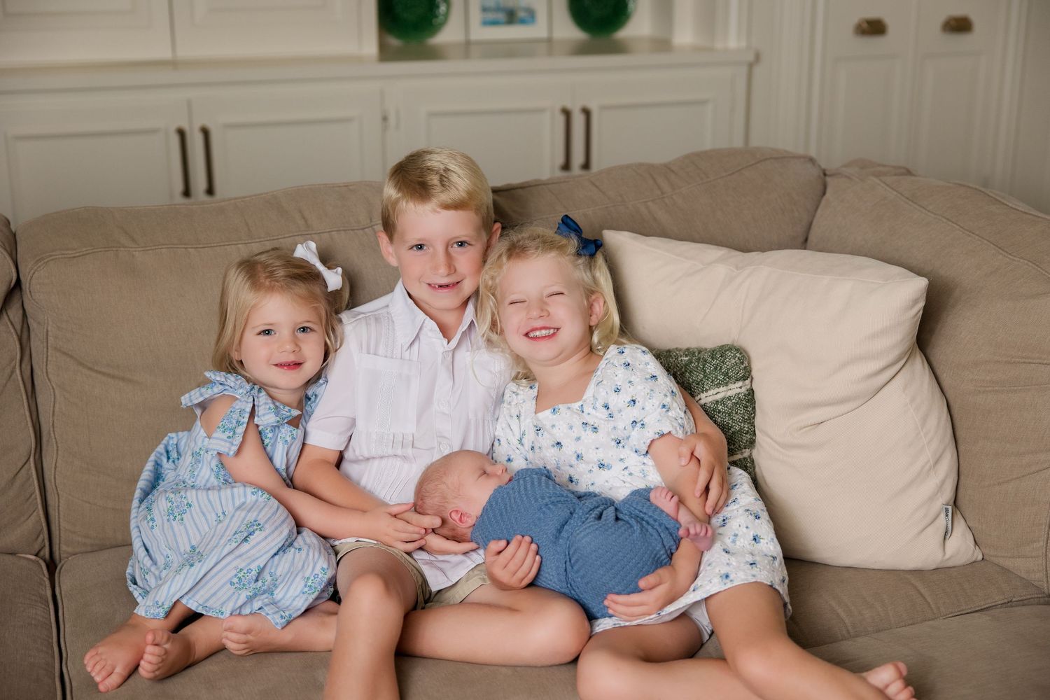 Children wearing light colored clothes sitting together on a beige couch in a living room.