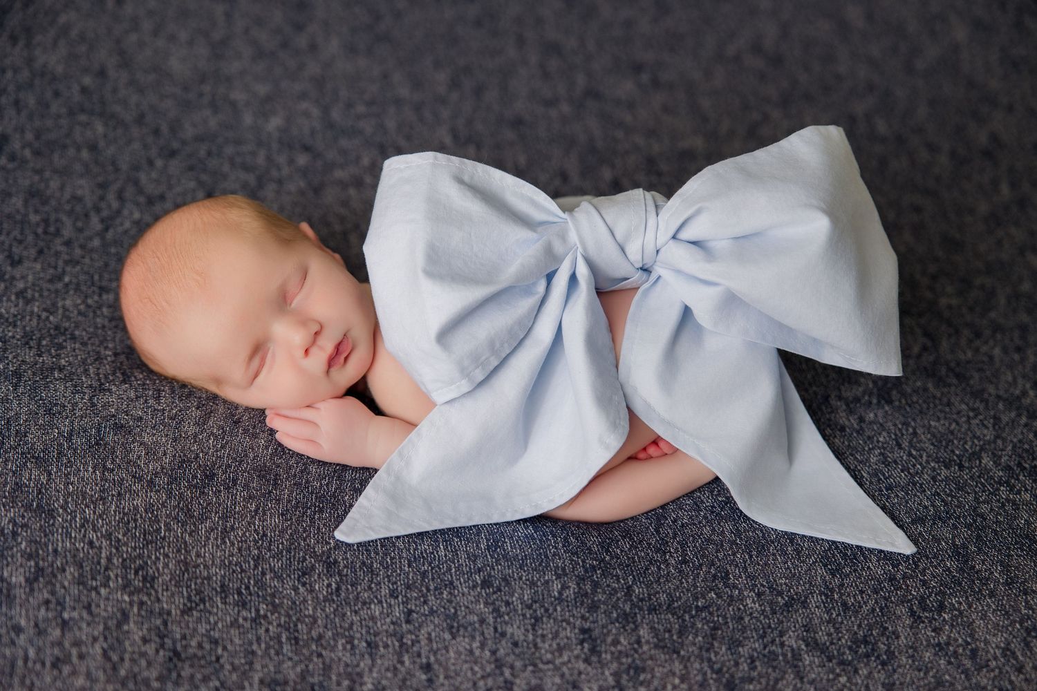 Sleeping newborn baby with white bow prop photographed on grey backdrop.