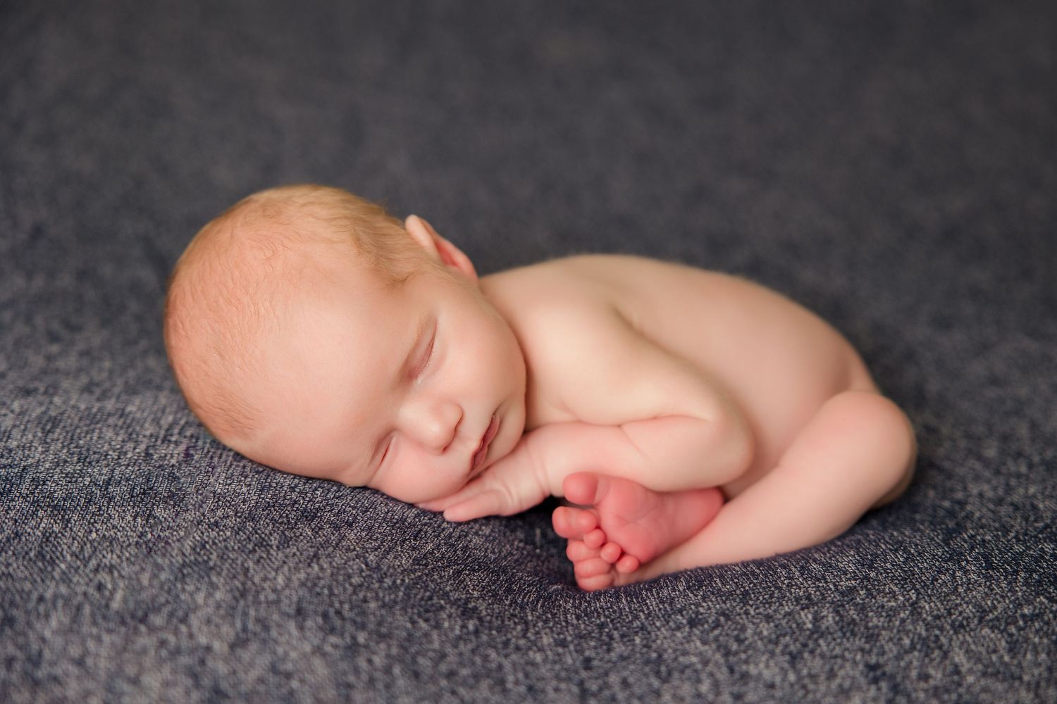 A sleeping newborn infant curled up peacefully on a soft grey blanket.