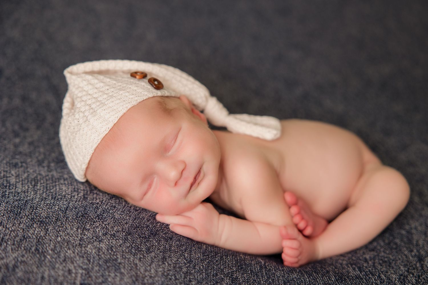 A sleeping newborn baby lies curled up on a dark gray blanket in a peaceful pose with feet visible.