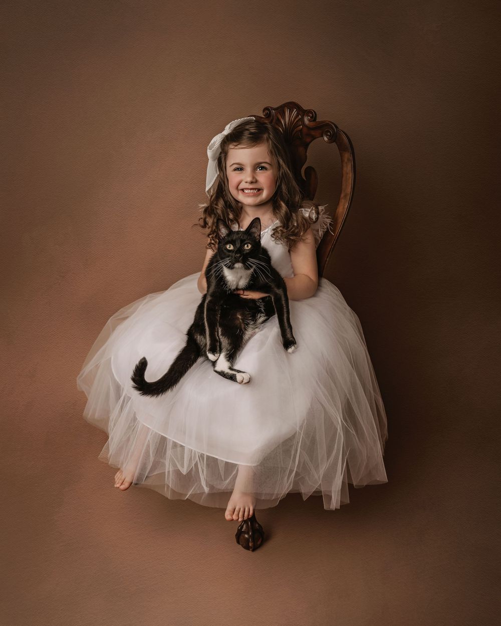 A portrait series showing a child in a white tulle dress holding a gray cat in different heartwarming poses against a brown backdrop.