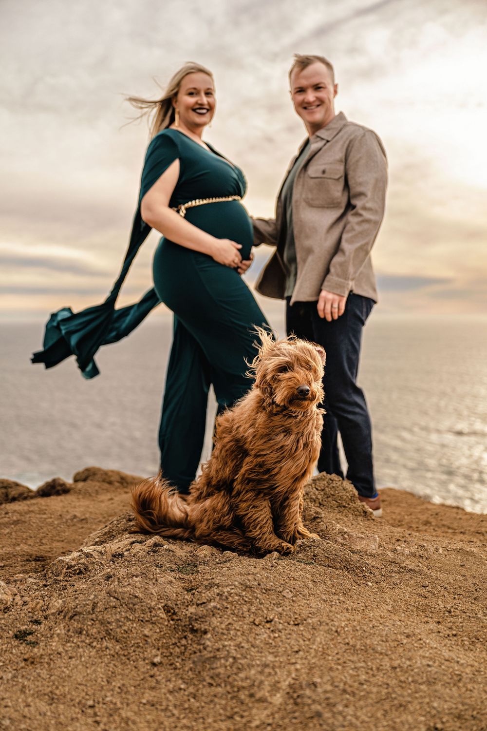 A couple stands at sunset on a beach cliff with their fluffy brown dog as part of their maternity photo session.