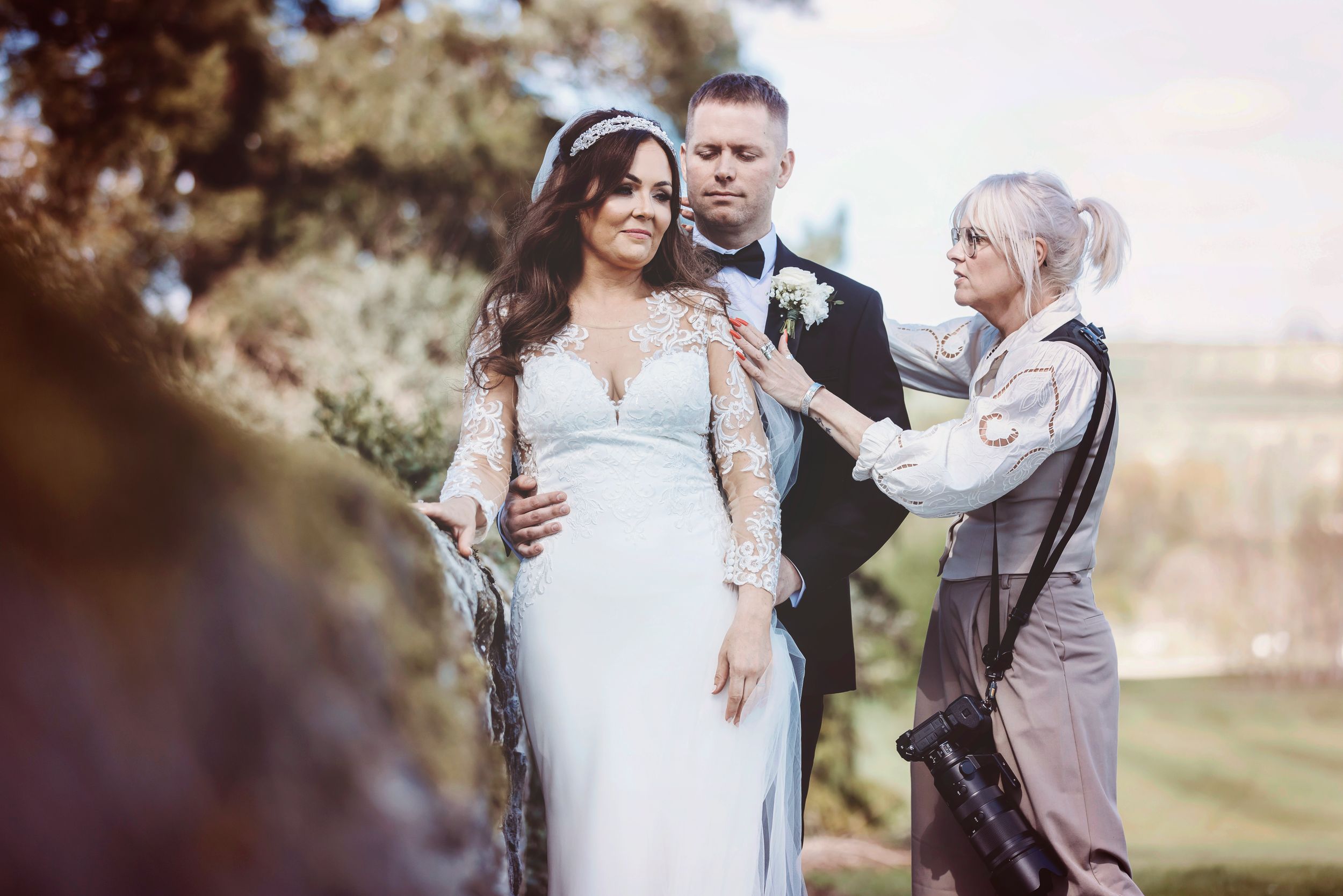 Professional photographer works with wedding couple outdoors during countryside photoshoot.