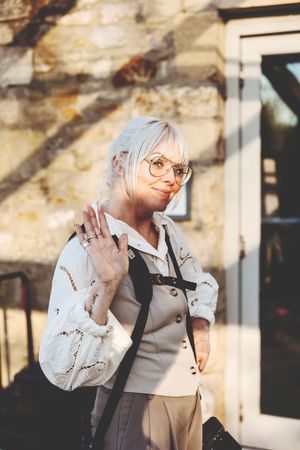 Photographer in glasses and light clothing waves while holding professional camera equipment outdoors.