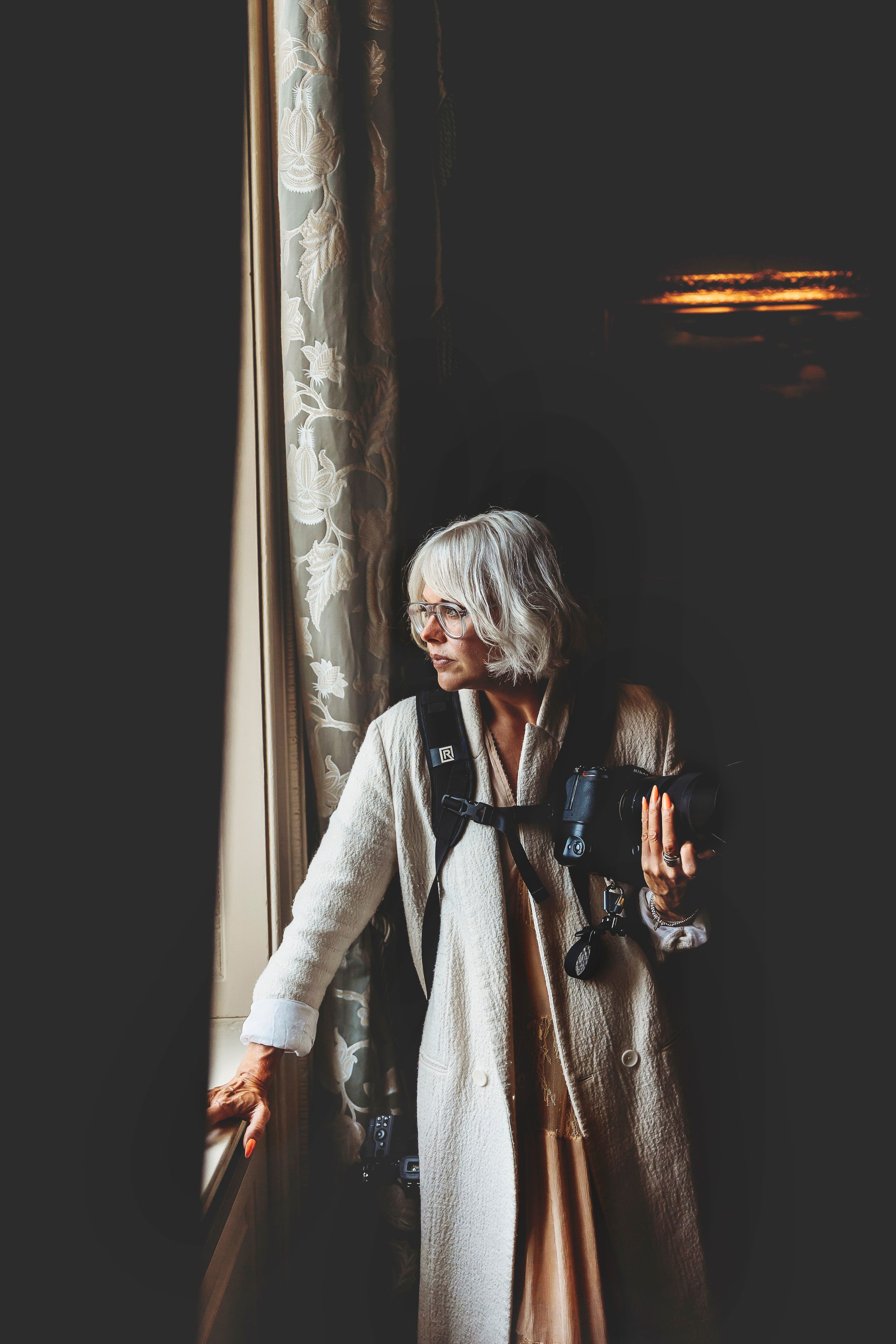 A figure in dramatic lighting stands pensively near a window wearing a long white coat and dark vest.