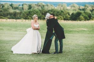 Wedding photographer captures candid moments between couple on grassy field with blue sky.