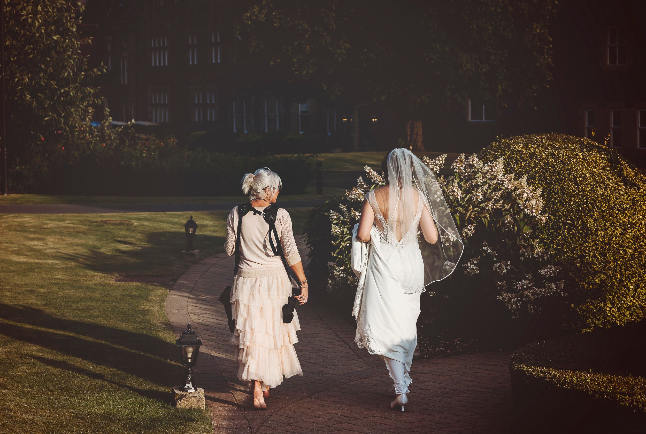 Couple walking along dark path at dusk with glowing lights in background.