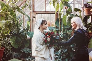 Professional photographer assists with bridal preparations in greenhouse filled with plants.