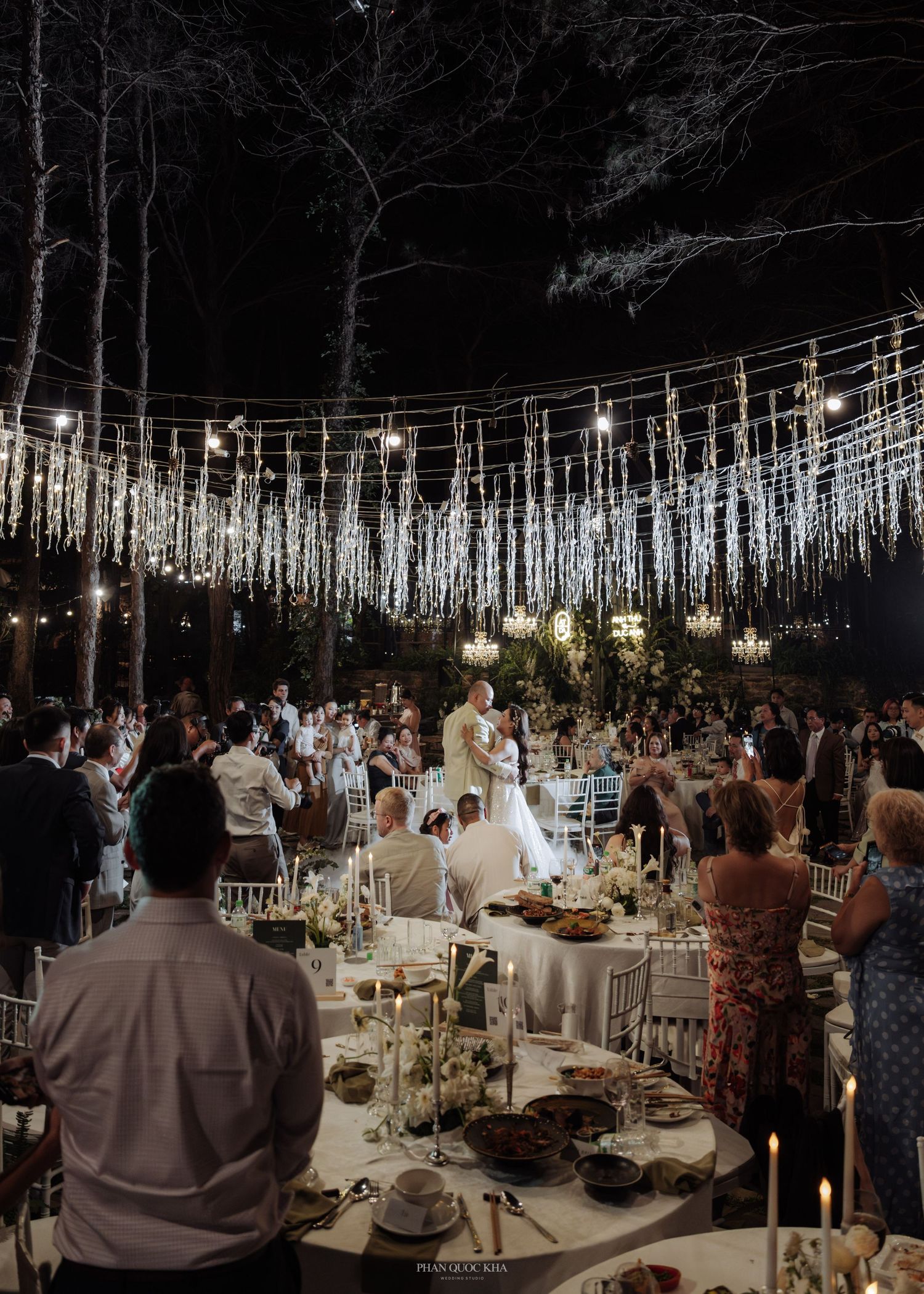 Elegant outdoor night wedding reception with string lights, chandeliers, and guests dining under the trees.