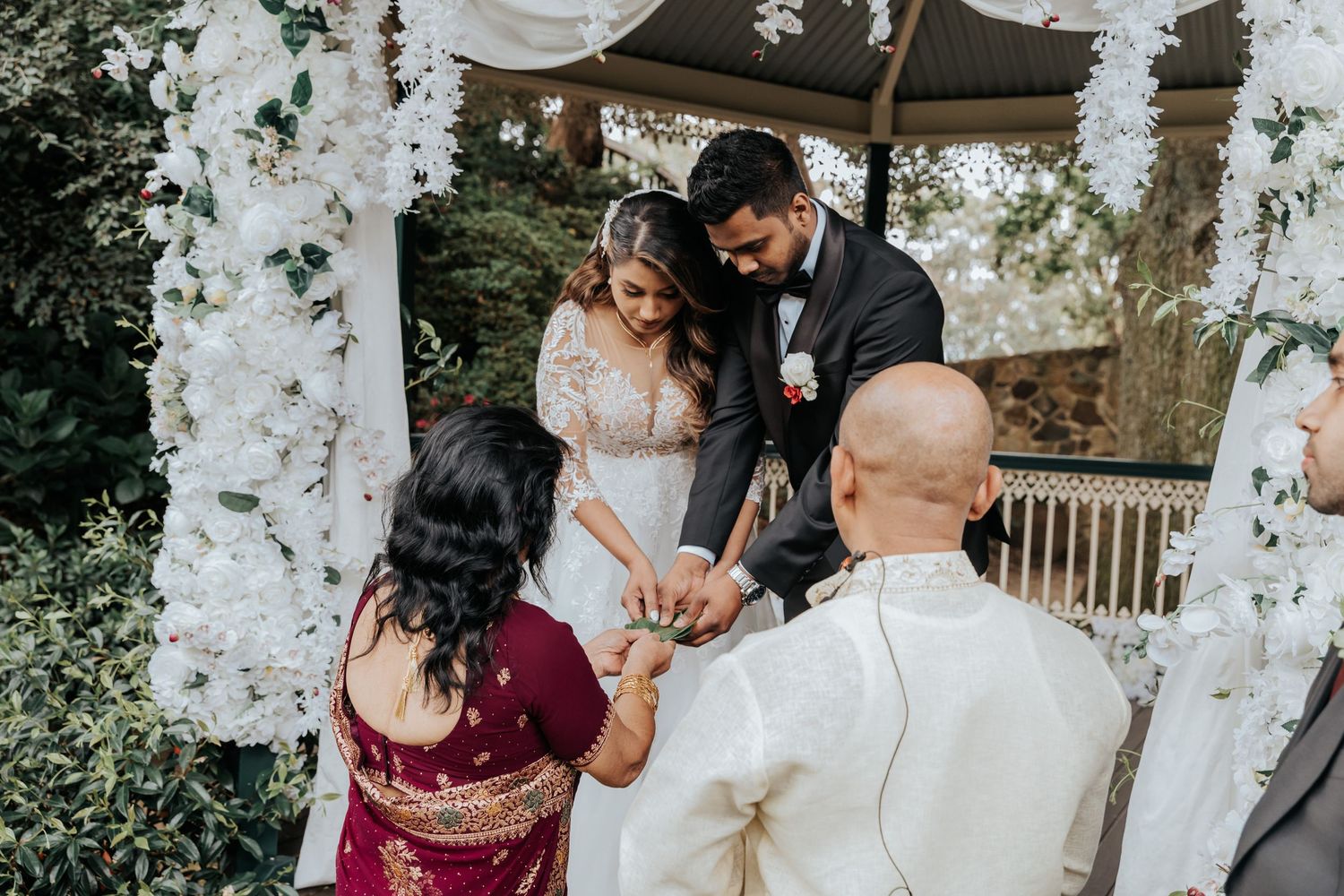A tender wedding ceremony moment captured under a floral archway with loved ones gathered at an outdoor gazebo.