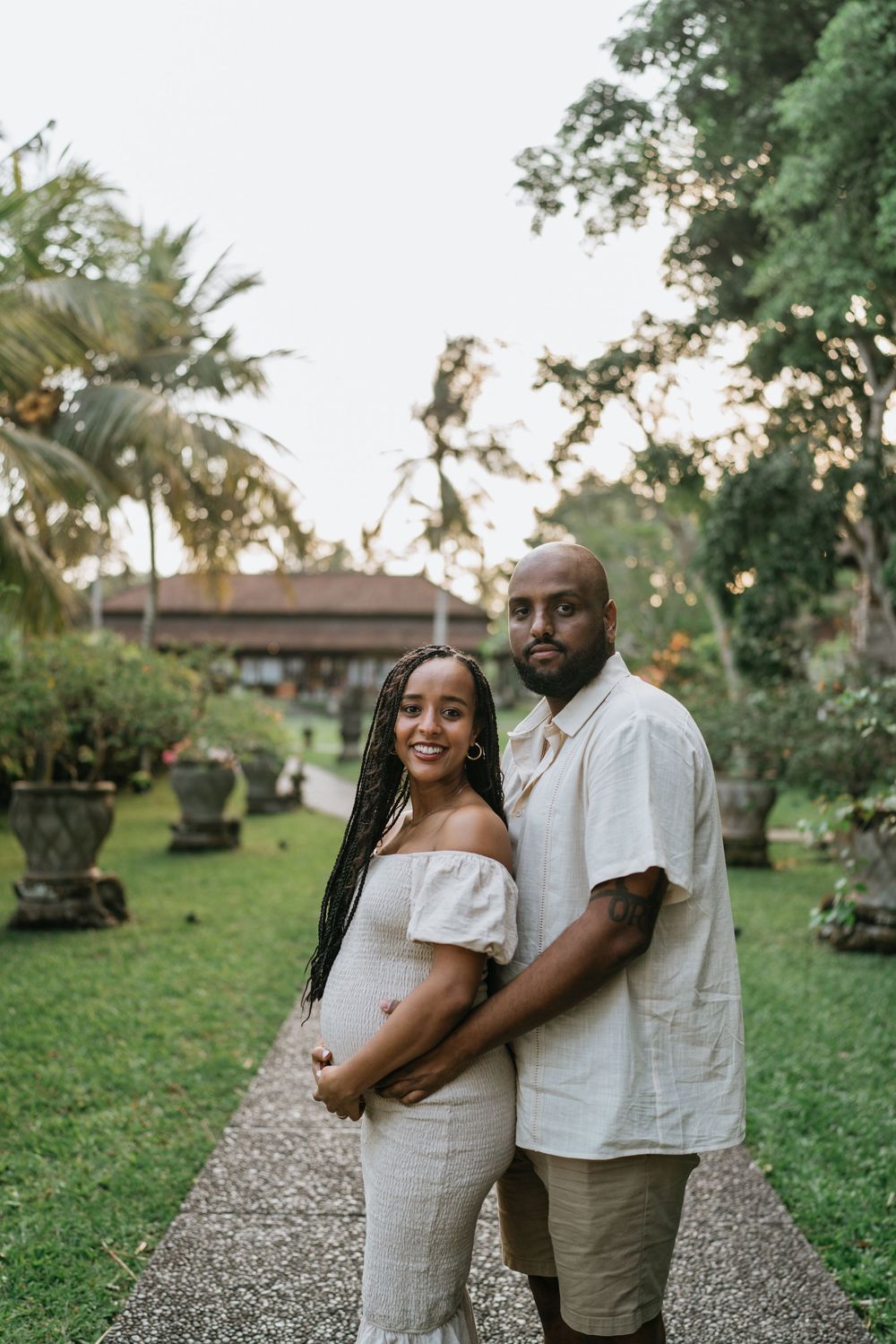 A couple in white outfits poses together on a gravel path surrounded by tropical gardens.