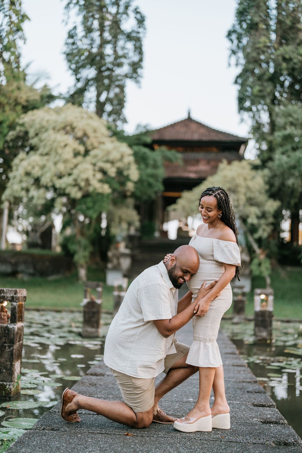 Couple poses for romantic photos by water with gazebo and trees in background.