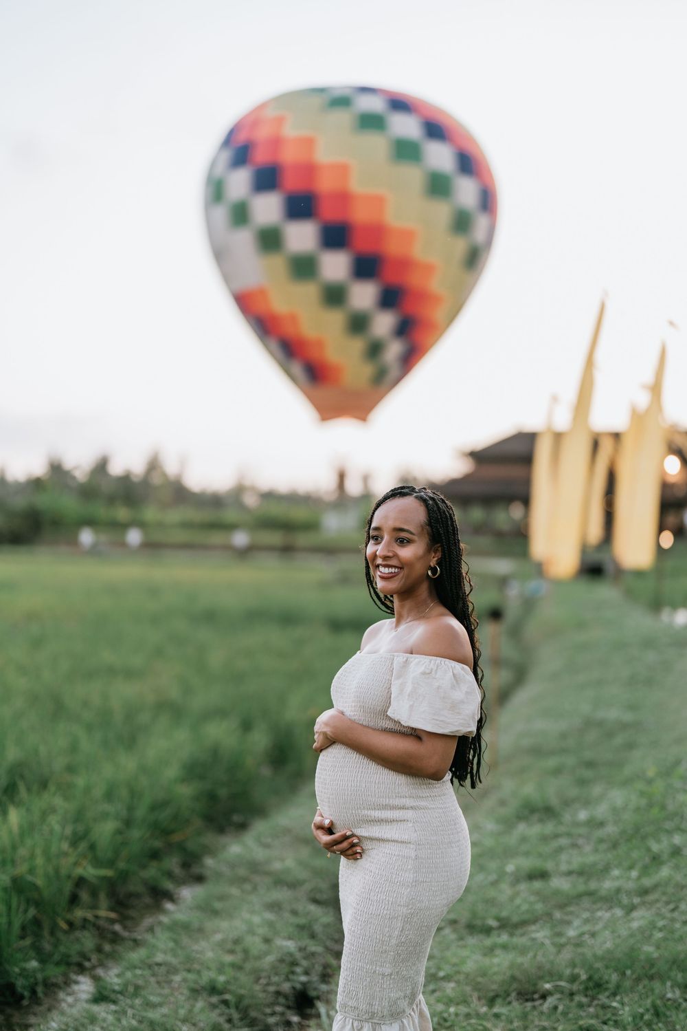 White dress contrasts beautifully with green field as hot air balloon floats in background.