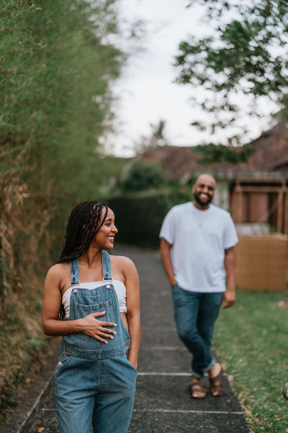 A couple walks along a scenic path wearing matching denim overalls in a casual outdoor setting.
