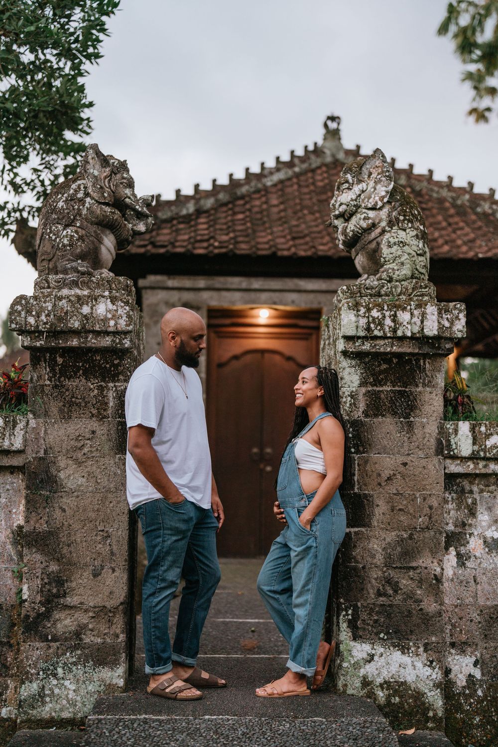 Couple stands together near an ornate stone wall and wooden door with traditional Balinese architecture.
