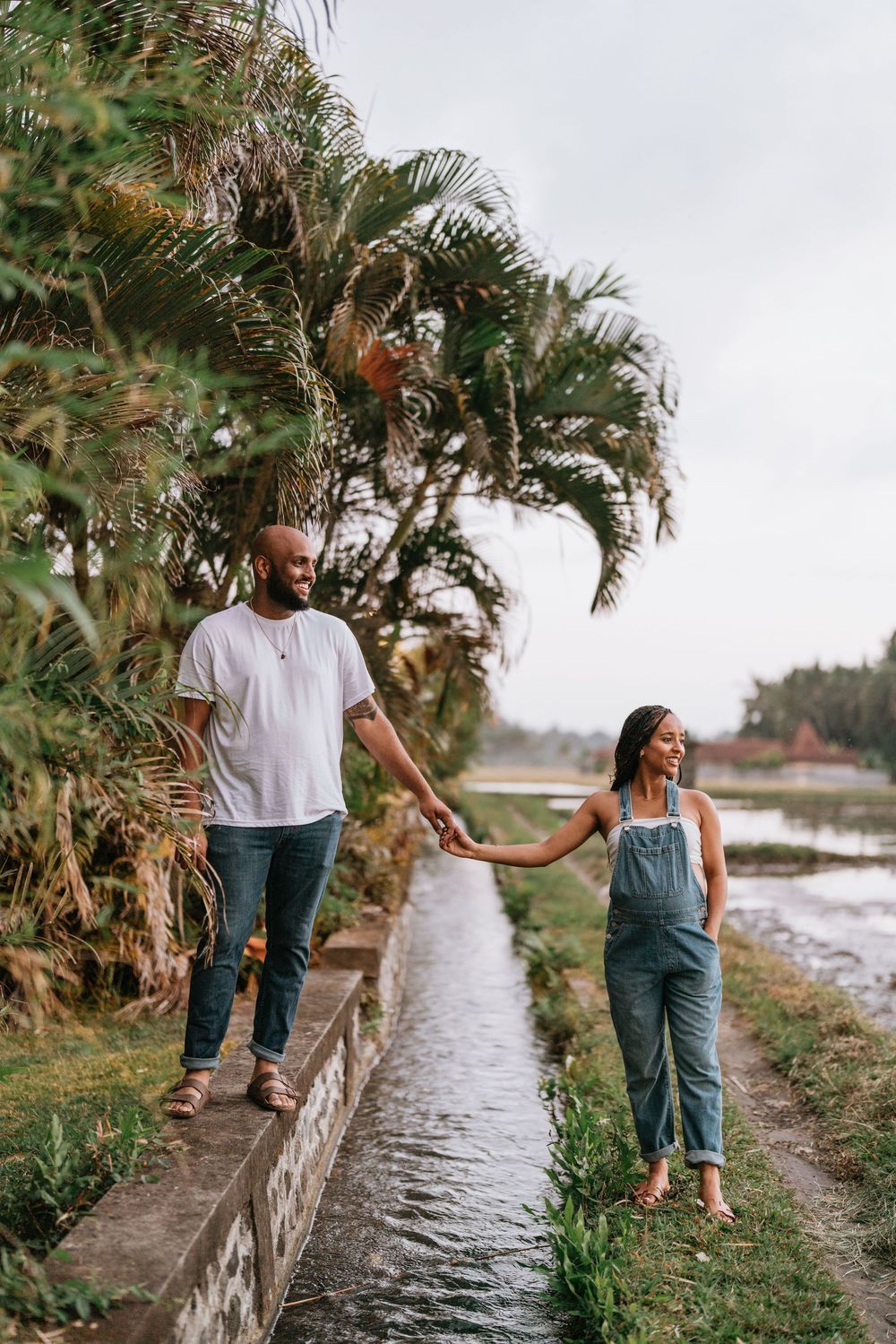 Couple walks along a waterfront path lined with palm trees during golden hour.