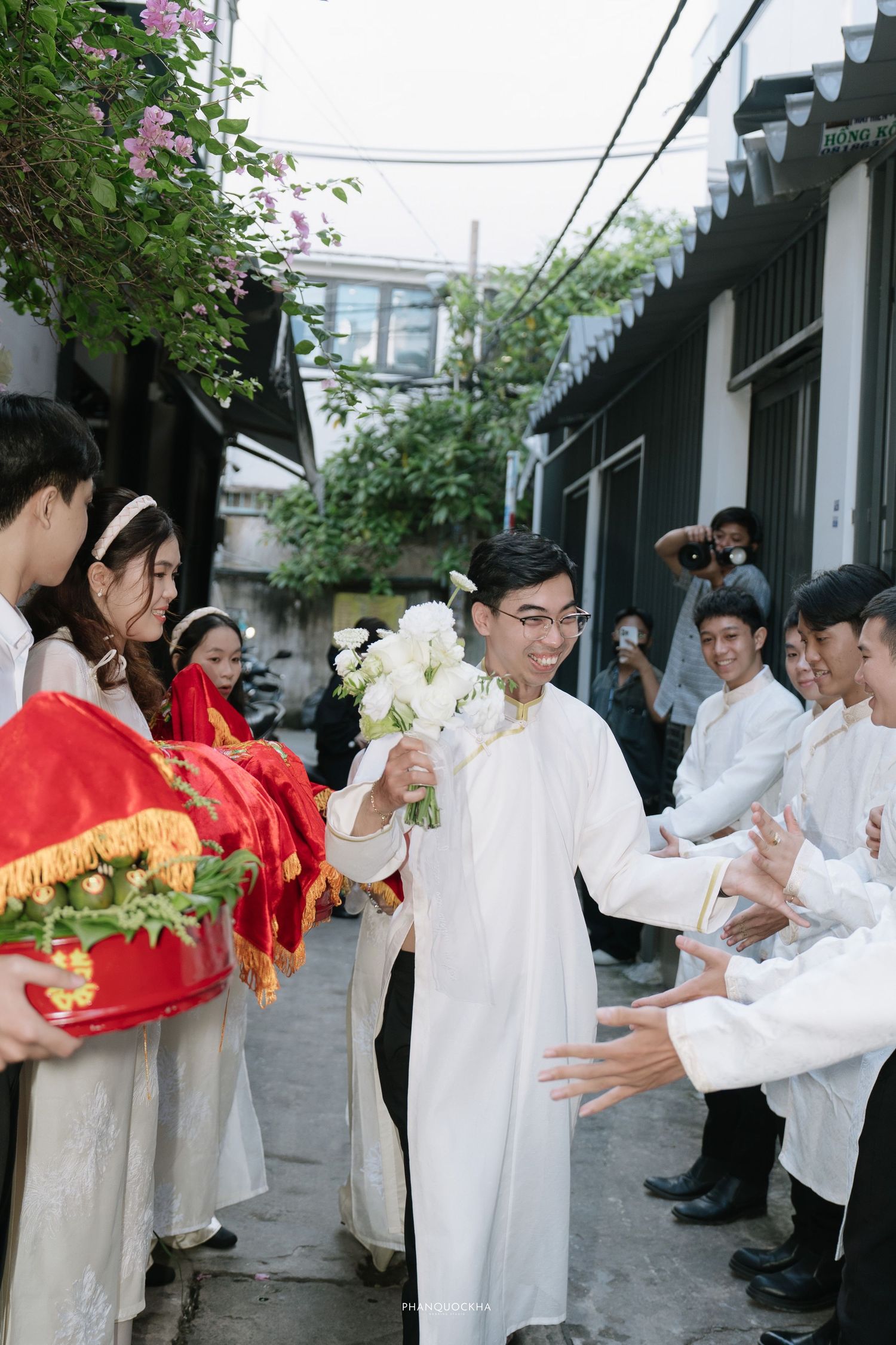 Groom in white ao dai holding white bouquet walks through alley lined with wedding party at Vietnamese ceremony.