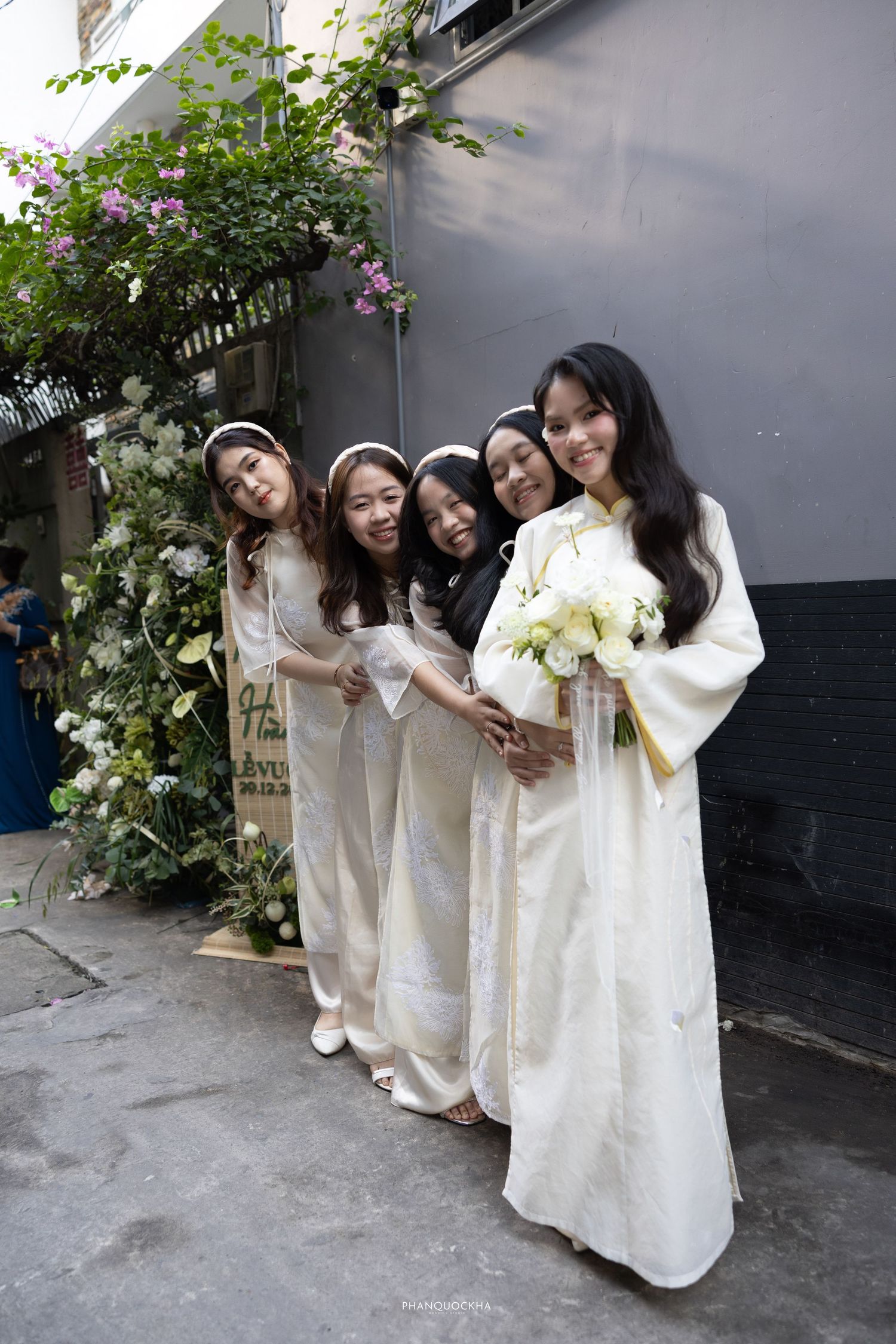Bride in white ao dai with bridesmaids posing playfully in alley with floral backdrop.