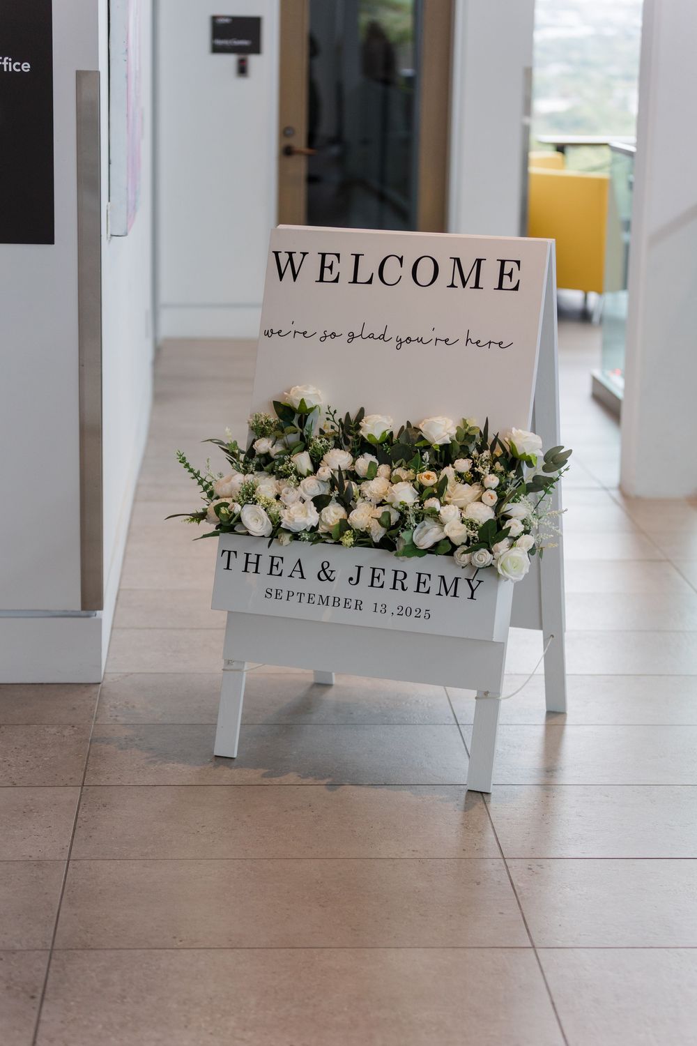 Wedding welcome signs with floral decorations displayed on white easels in a light-filled venue.