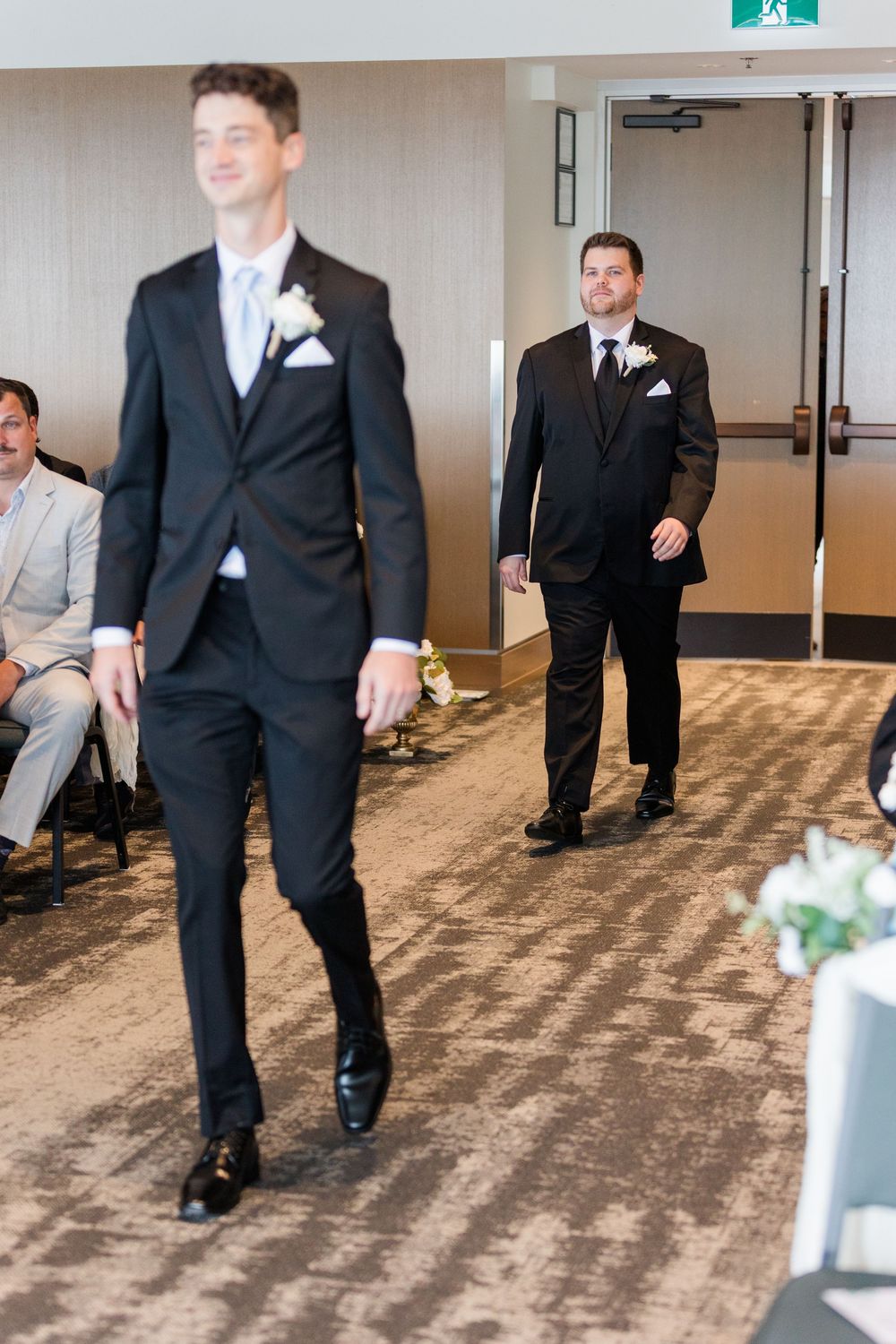 Groomsmen in classic black tuxedos walk down the aisle at an indoor wedding ceremony.