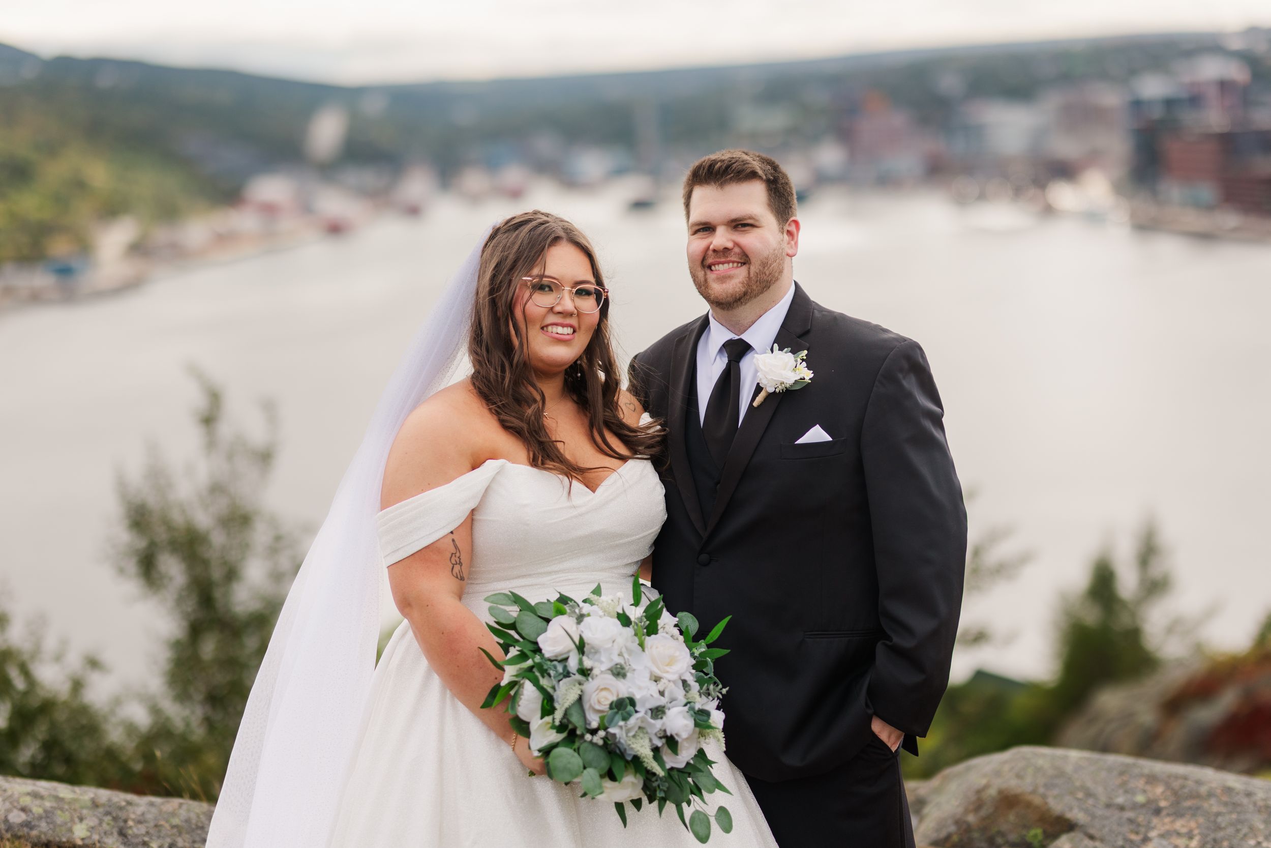 A wedding couple stands on a scenic coastal overlook holding a white and green floral bouquet.