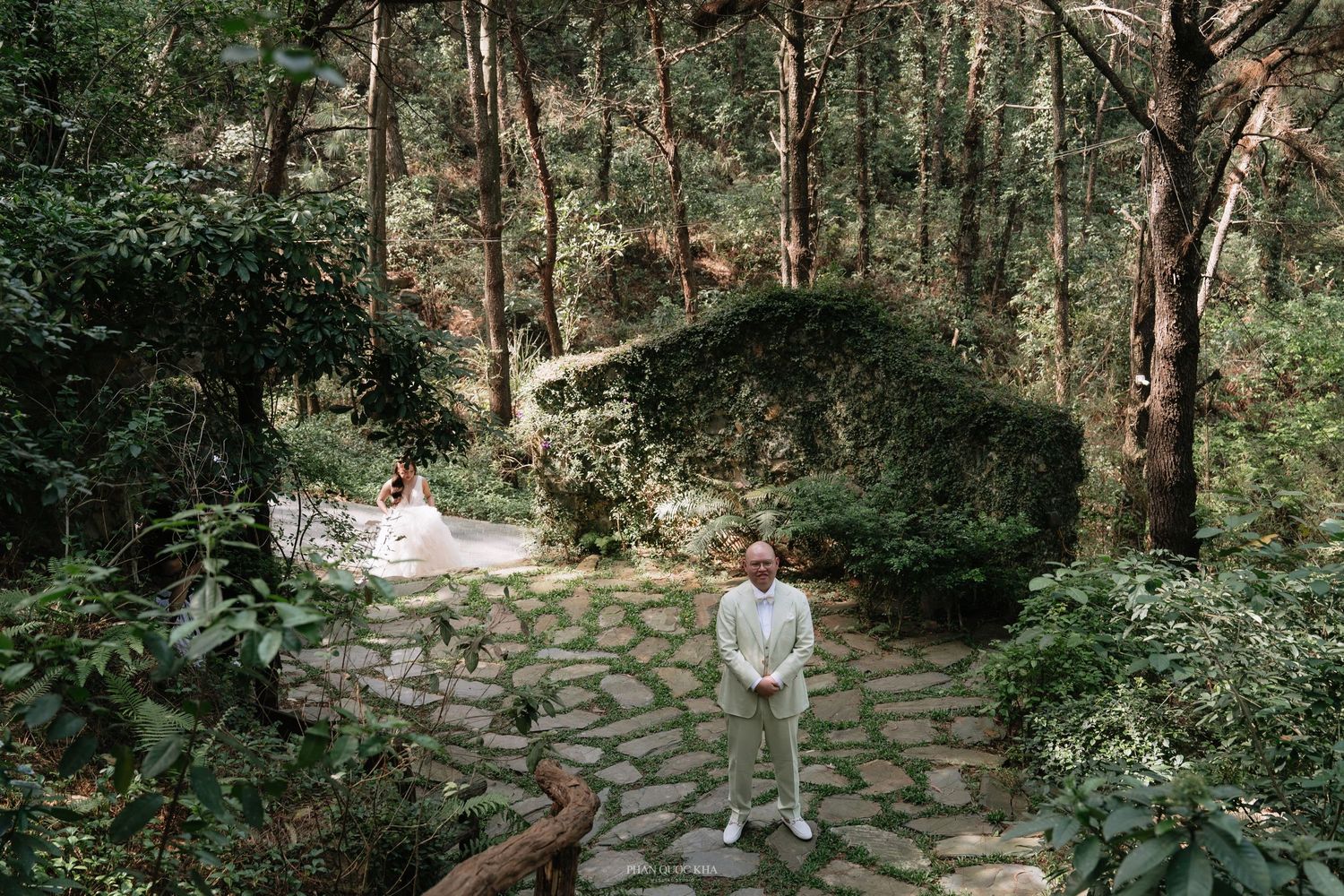 Bride and groom standing apart on a stone path surrounded by a serene forest setting.