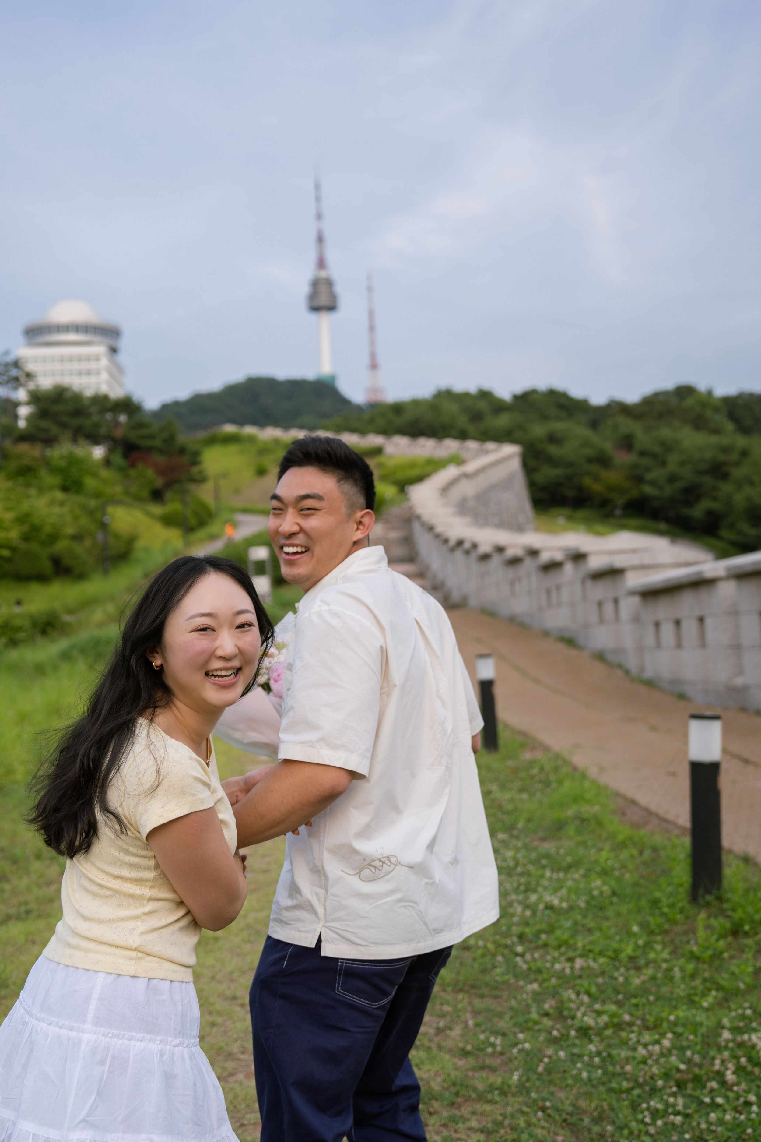 Couple sharing playful moments on a hillside path with Seoul Tower in the background.