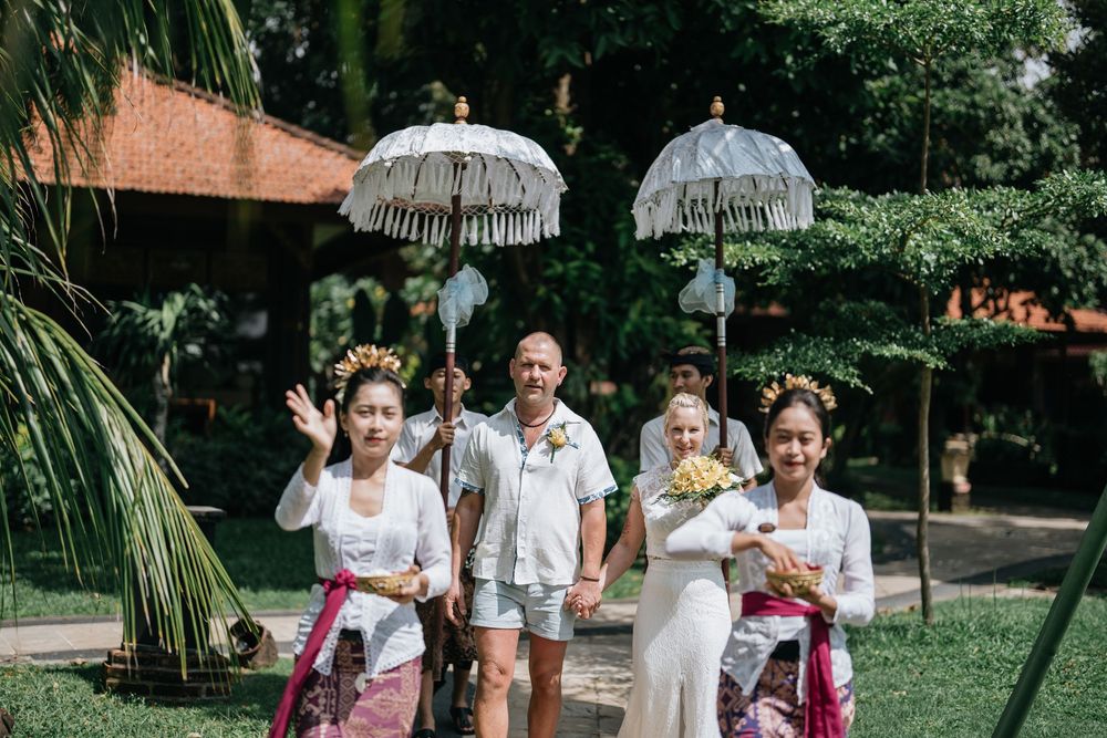 Bridal party in white dresses carrying parasols while walking through a sunny garden.