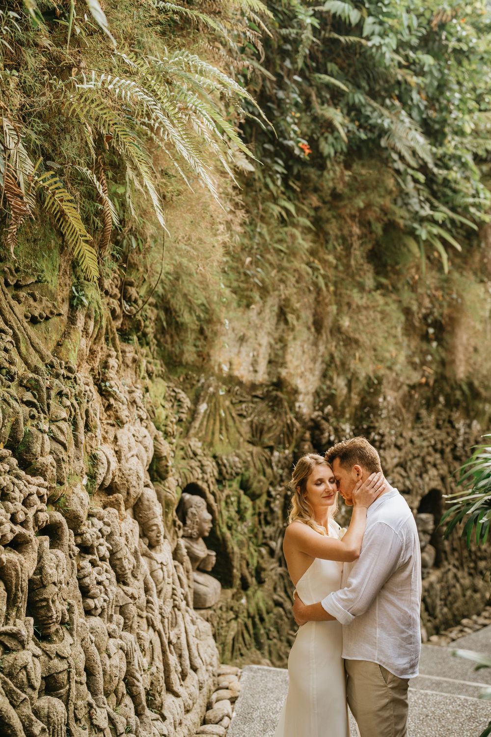 Wedding couple embracing by a stone wall path surrounded by tropical greenery.