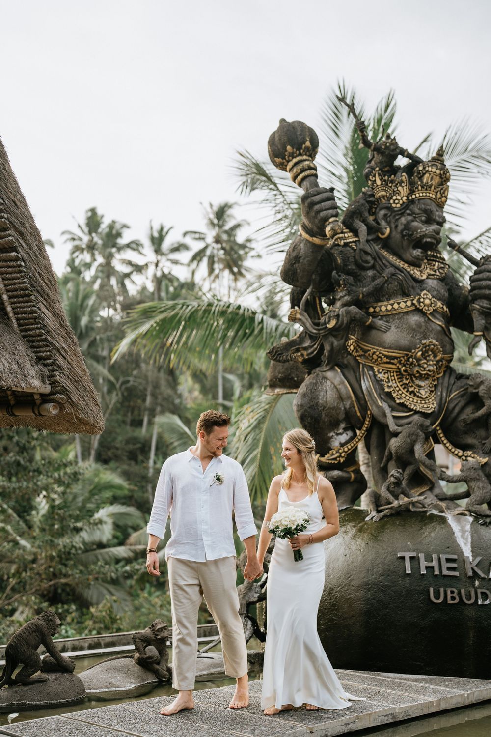 Wedding couple posing near traditional Balinese temple statues and palm trees.