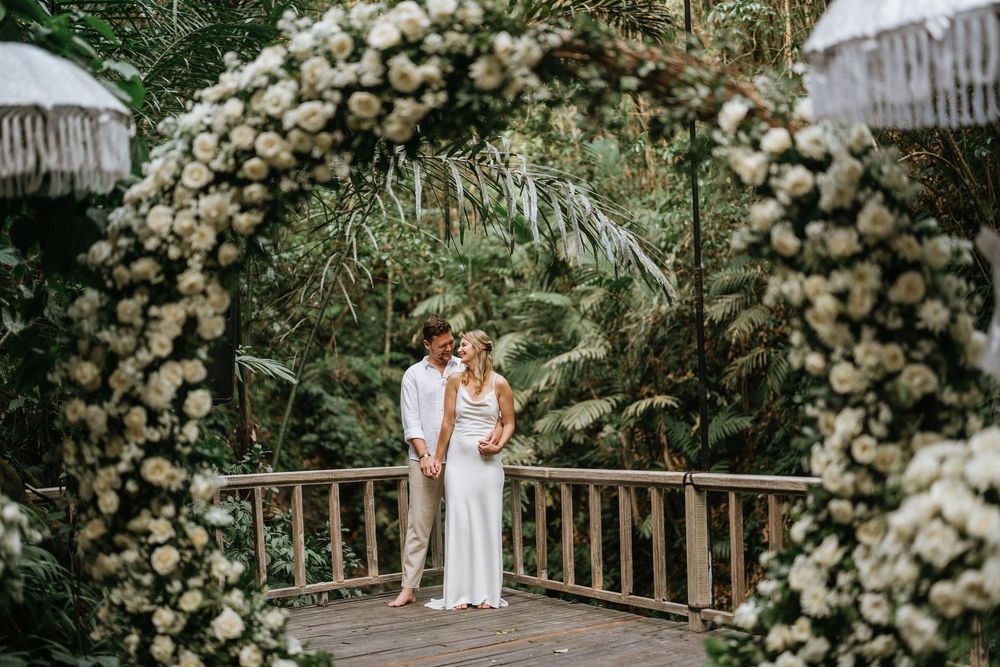 Romantic garden wedding ceremony setting with white floral arch and wooden chairs under string lights.