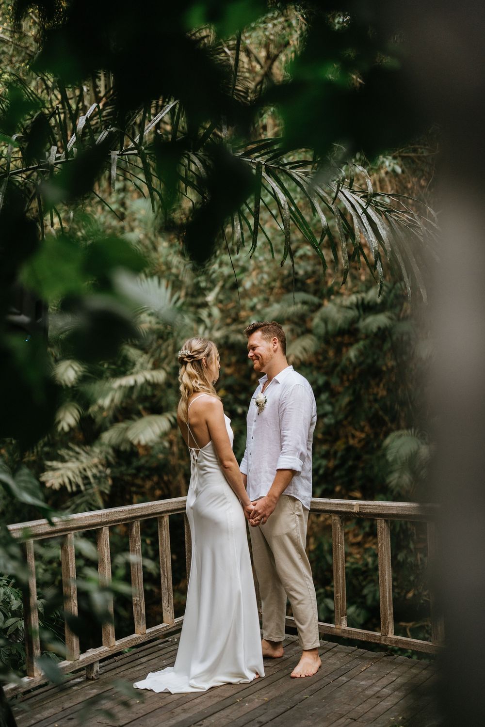 Couple shares intimate moment on wooden deck surrounded by lush tropical garden and white flowers.