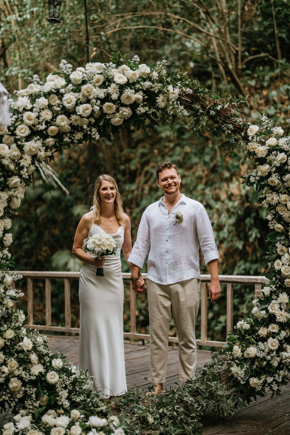 Wedding couple walks along wooden pathway adorned with cascading white roses and greenery.