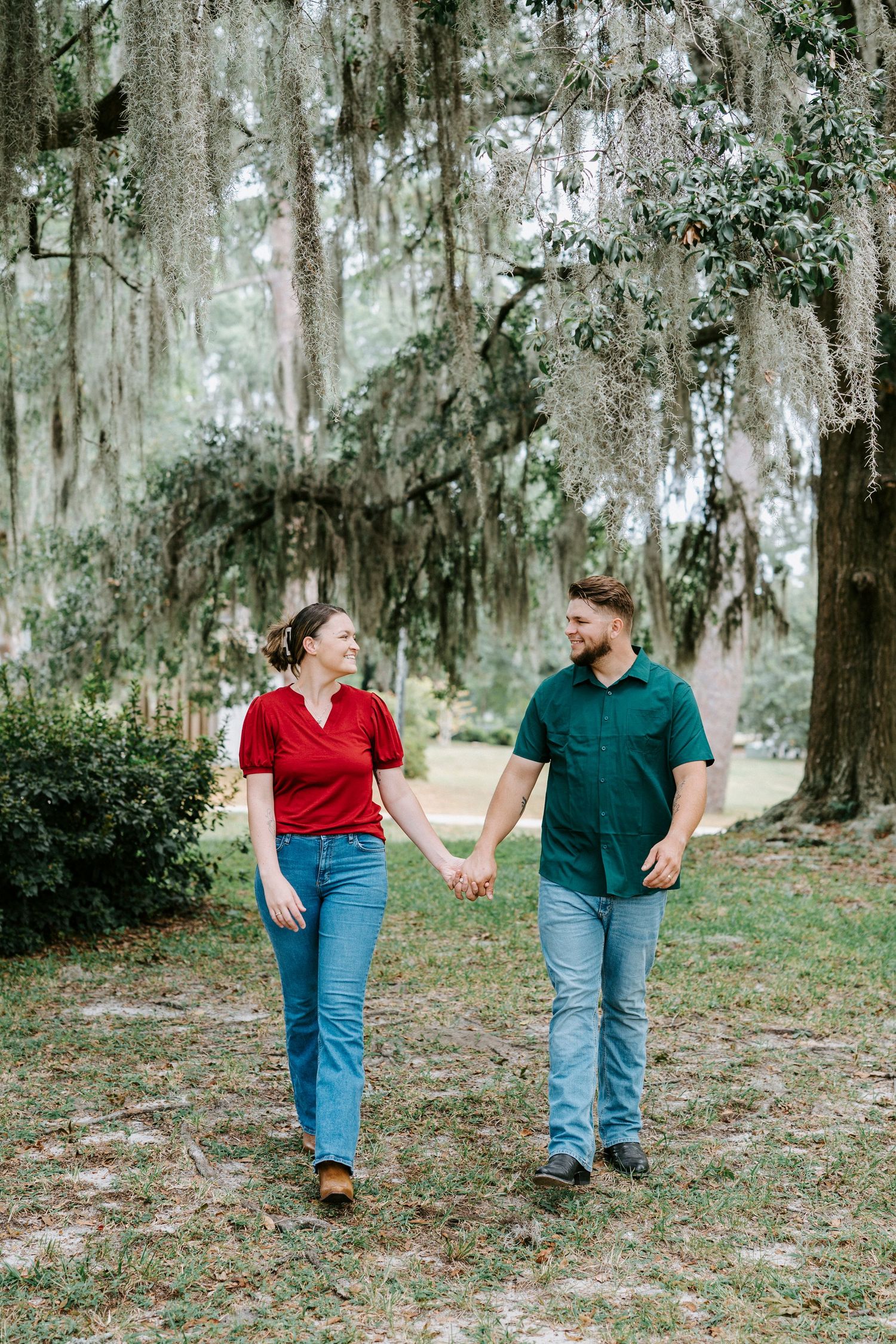 A couple walking hand in hand through a scenic moss-covered oak tree forest in casual red and green attire.