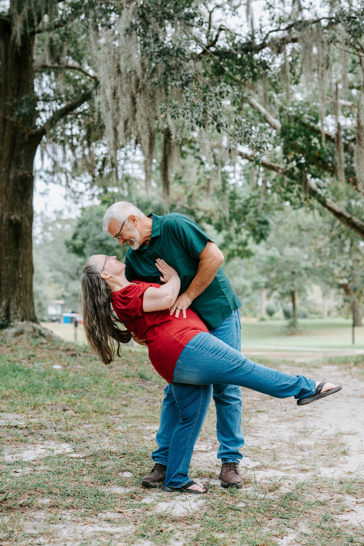 A couple in casual clothing sharing a romantic dip pose together in a wooded outdoor setting.