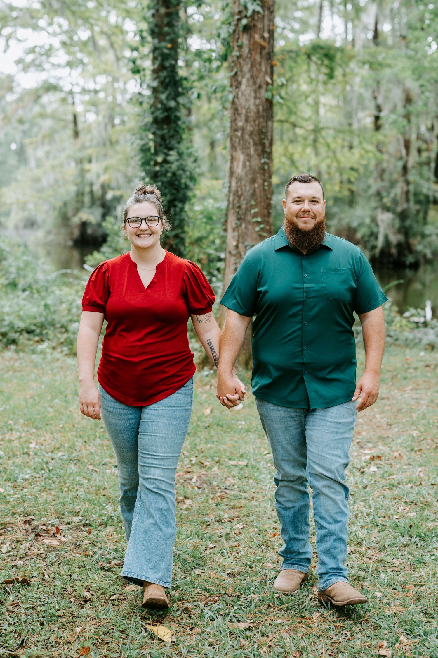 A couple wearing red and green shirts walk hand in hand through a wooded outdoor setting.