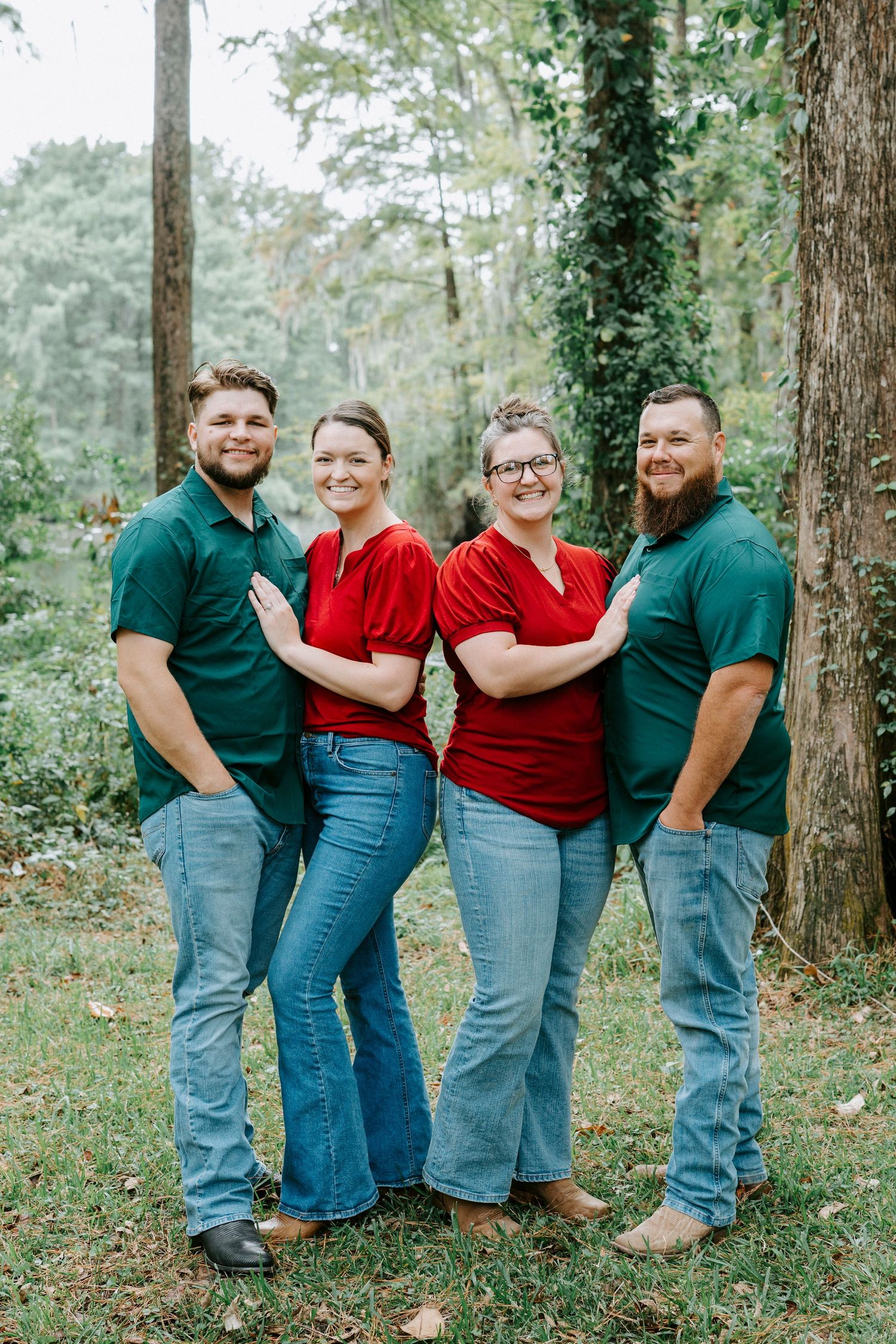 Two couples in coordinating red and green tops pose together outdoors in a wooded setting.