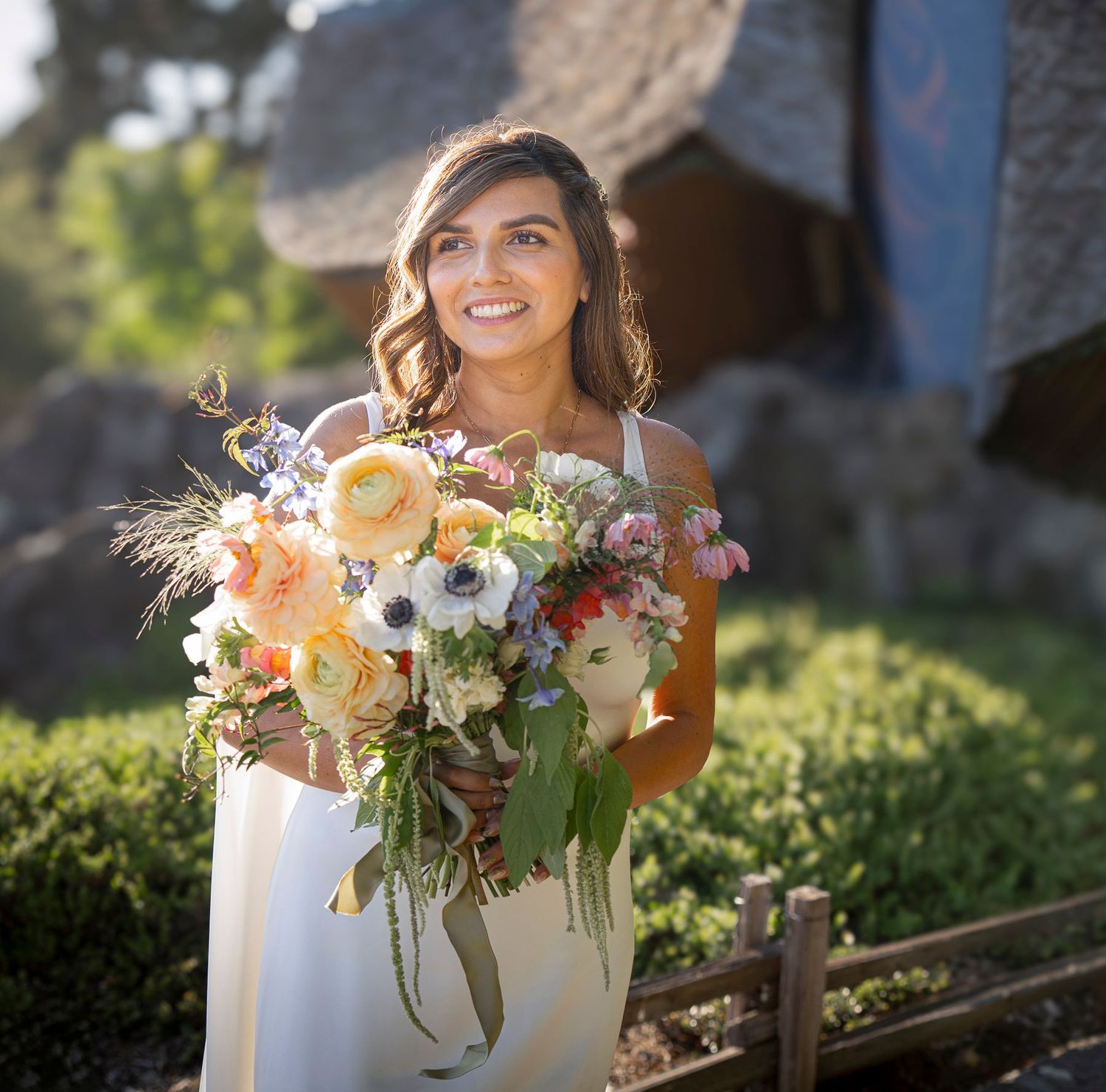 A bride in front of The Sea Ranch Chapel in golden sunlight holds a lush bouquet of peach and purple wildflowers