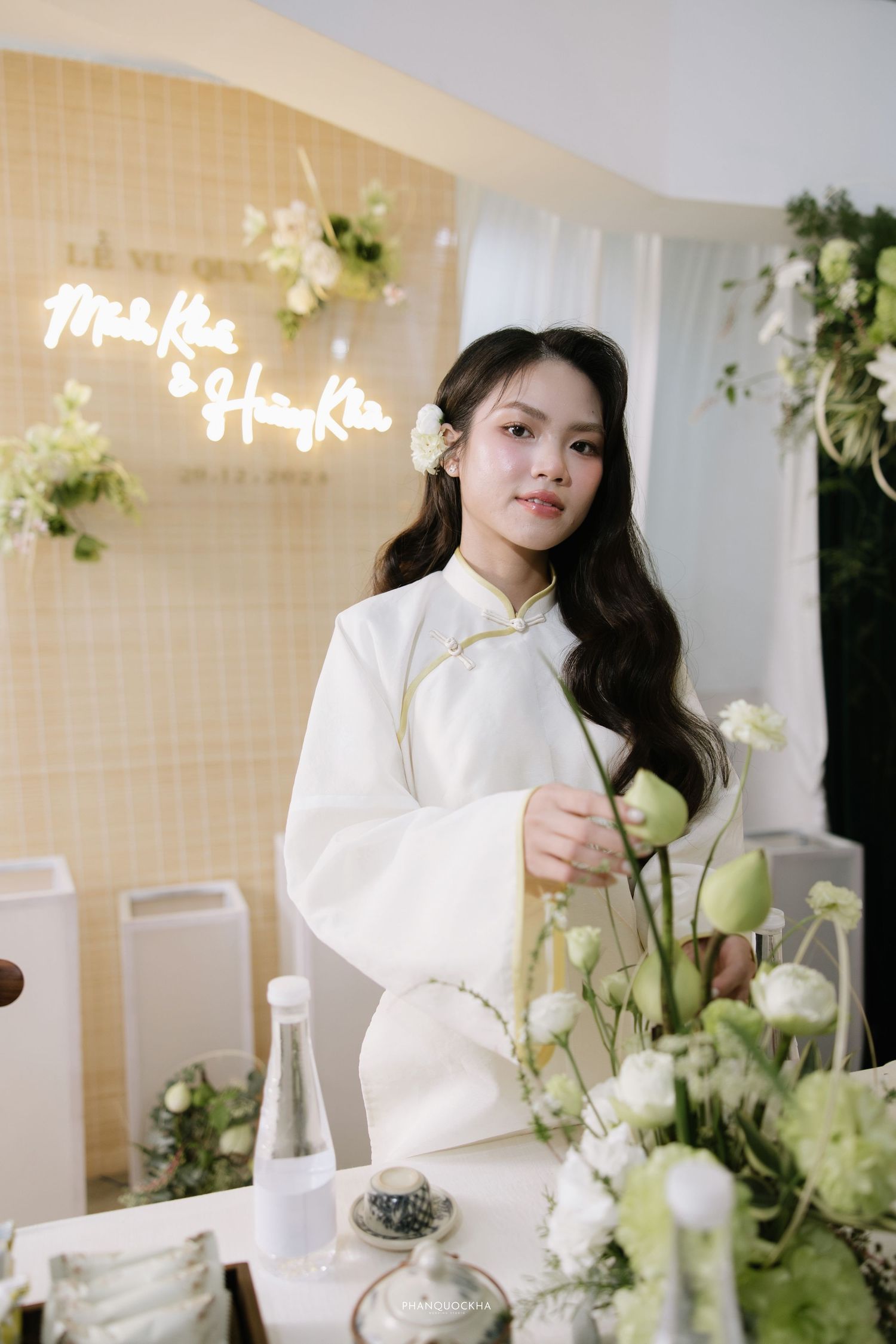 Elegant woman in white traditional ao dai stands beside white and green floral arrangement at wedding.