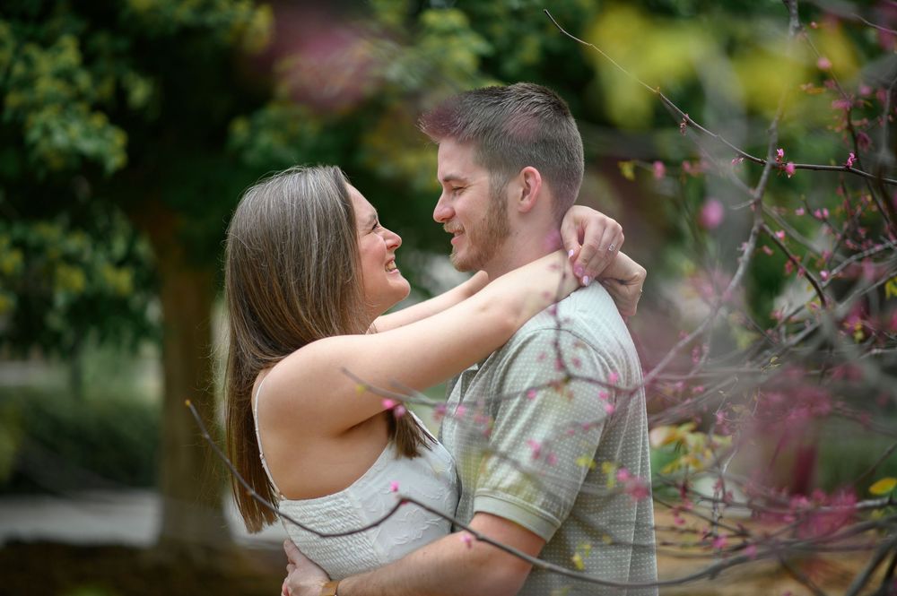Couple embracing in a whimsical garden setting with soft natural lighting and blurred floral elements.