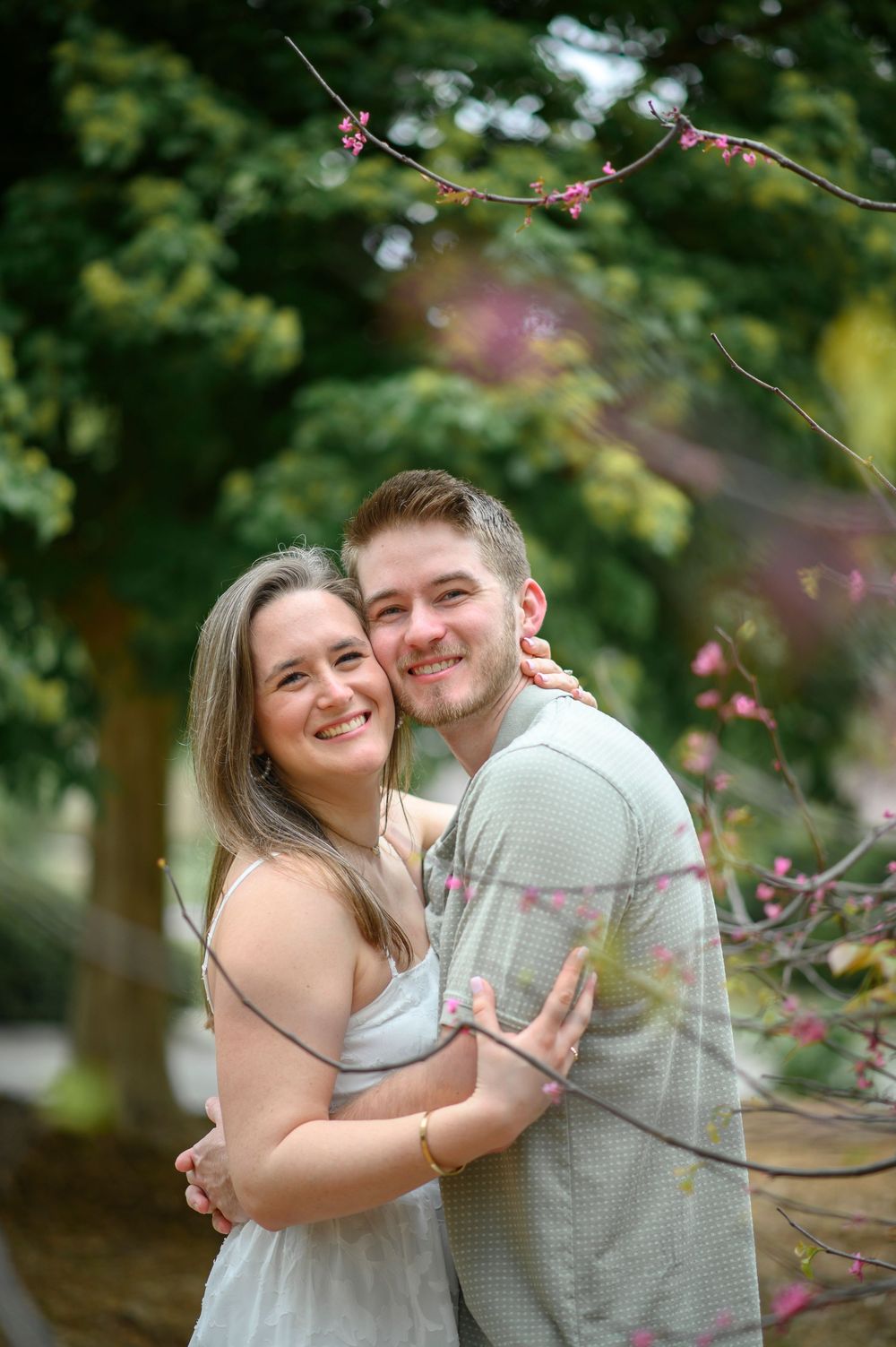 A couple embraces and smiles together outdoors with blooming spring branches in the background.