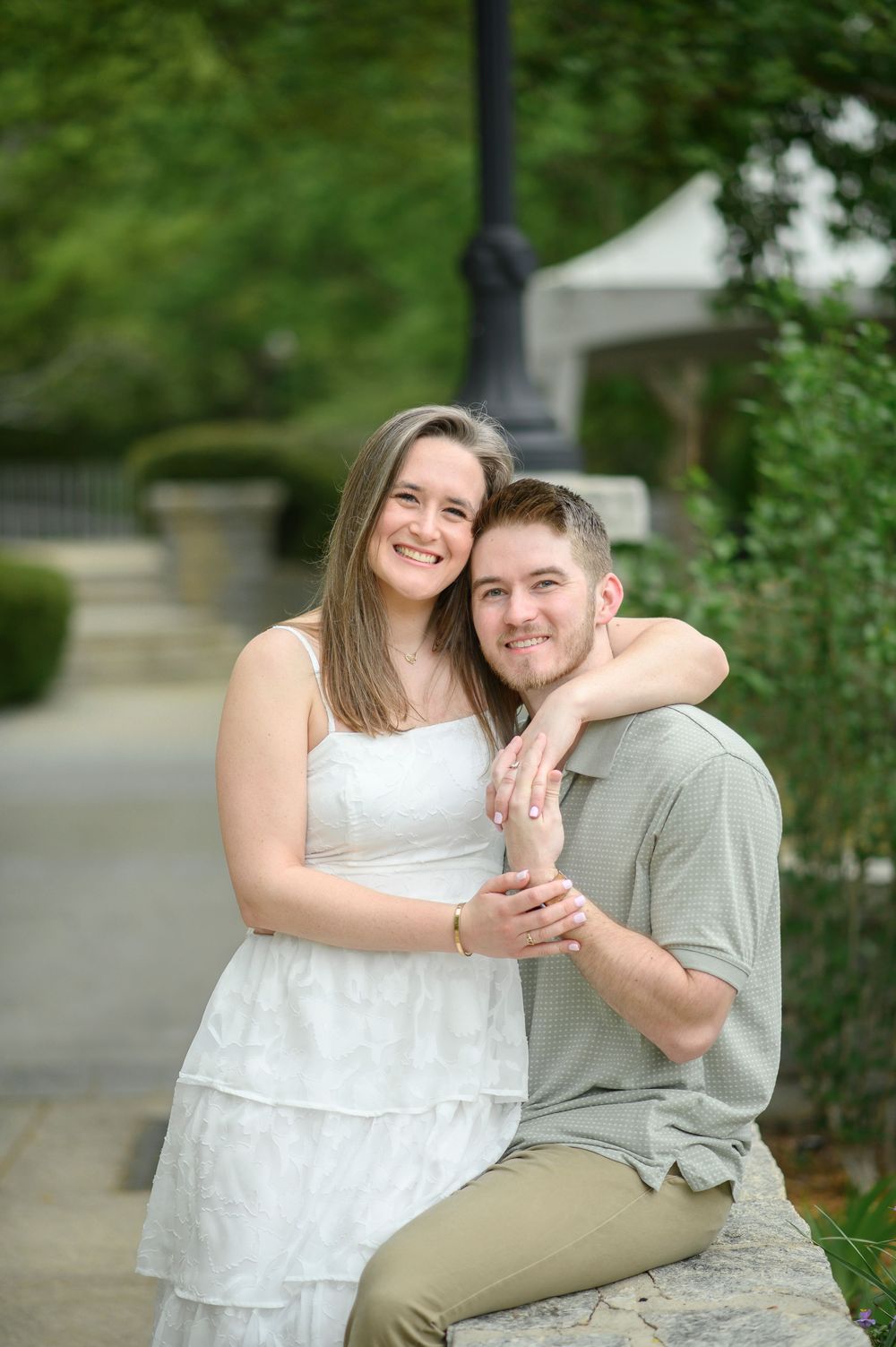 A couple sits together on a stone wall in a garden setting, embracing and smiling on a sunny afternoon.