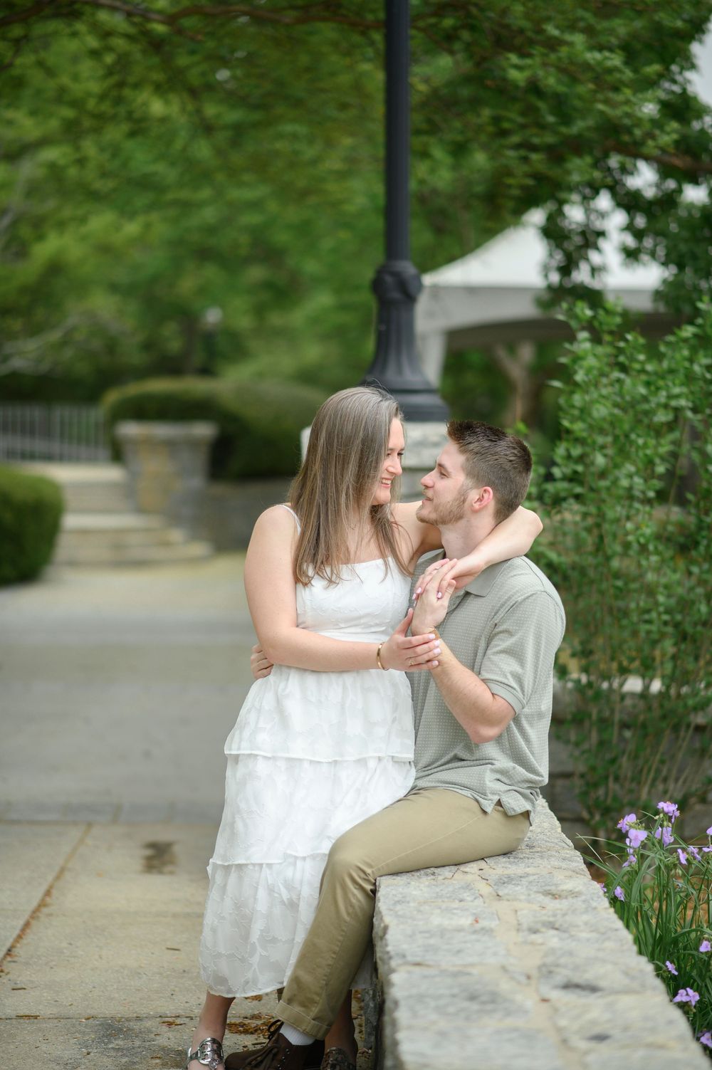 Romantic scene of couple sitting together on stone ledge surrounded by blooming flowers and greenery.