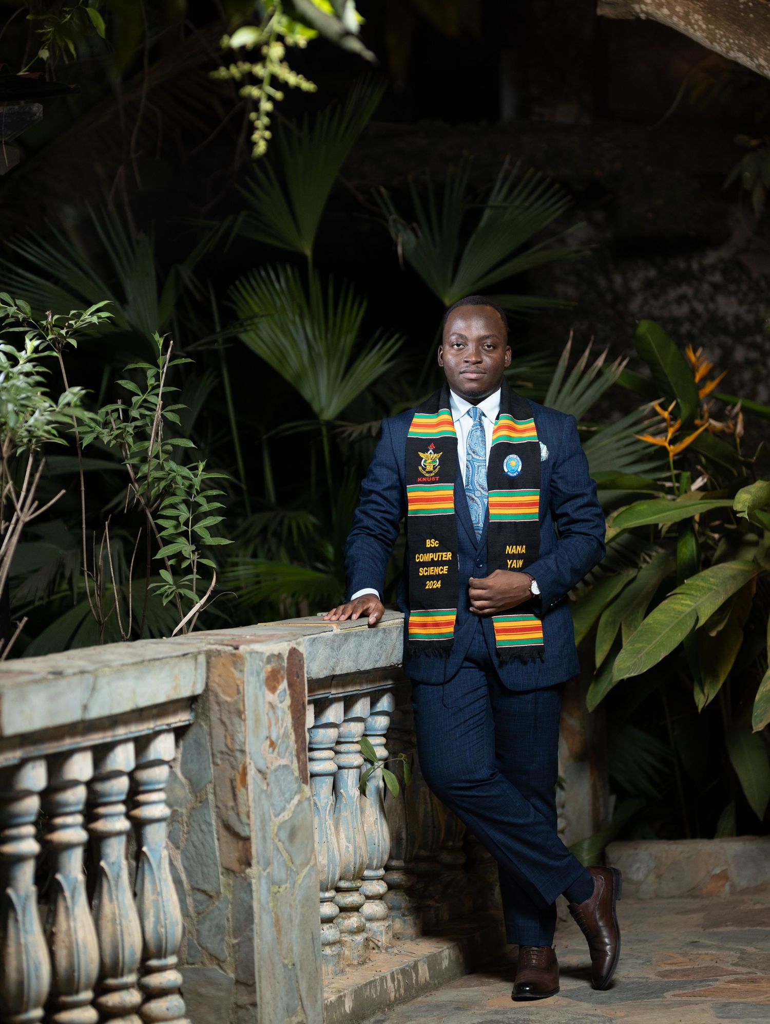 Graduate wearing academic regalia poses against wooden railing in modern interior space with dramatic lighting.
